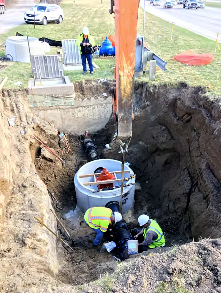 Excavator lowering a precast manhole structure while the crew guides it into place